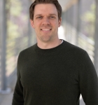 Erik's headshot, where he stands on a bridge overlooking a sunny forest and wears a black, long-sleeved shirt.