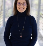 Sophie's headshot, where she stands on a bridge overlooking a forest and wears a black turtleneck and a long necklace. She has shoulder-length brown hair and glasses.