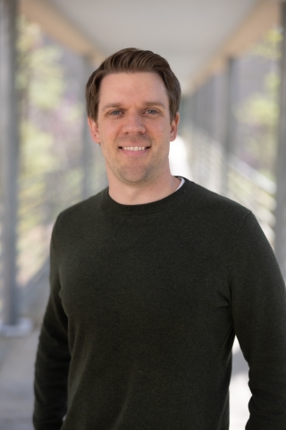 Erik's headshot, where he stands on a bridge overlooking a sunny forest and wears a black, long-sleeved shirt.