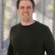 Erik's headshot, where he stands on a bridge overlooking a sunny forest and wears a black, long-sleeved shirt.