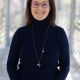 Sophie's headshot, where she stands on a bridge overlooking a forest and wears a black turtleneck and a long necklace. She has shoulder-length brown hair and glasses.